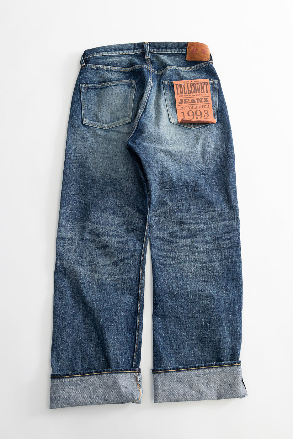 Flat lay of hard-washed indigo denim jeans, back view, showing rear pockets and leather patch.