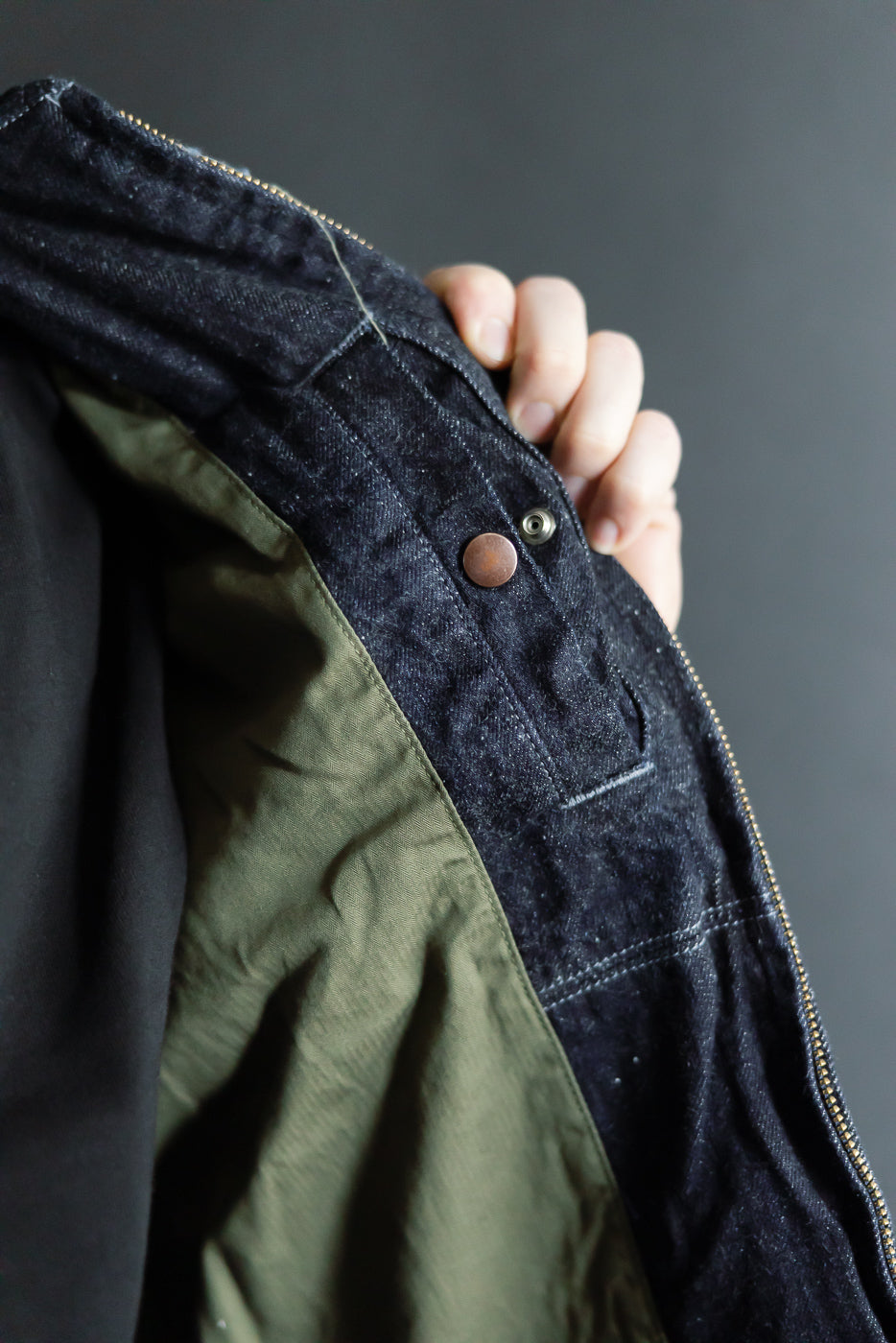 Close-up of dark denim jacket's front placket with copper button, olive lining, and zipper detail