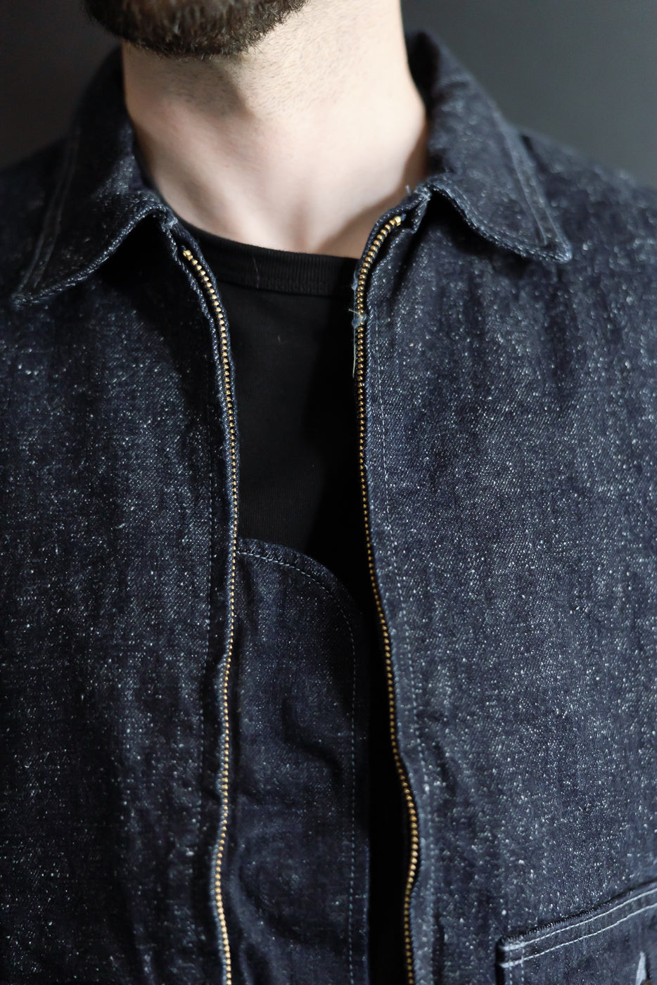 Close-up front view of male model in dark denim jacket with gold zipper, collar, and chest pocket detail