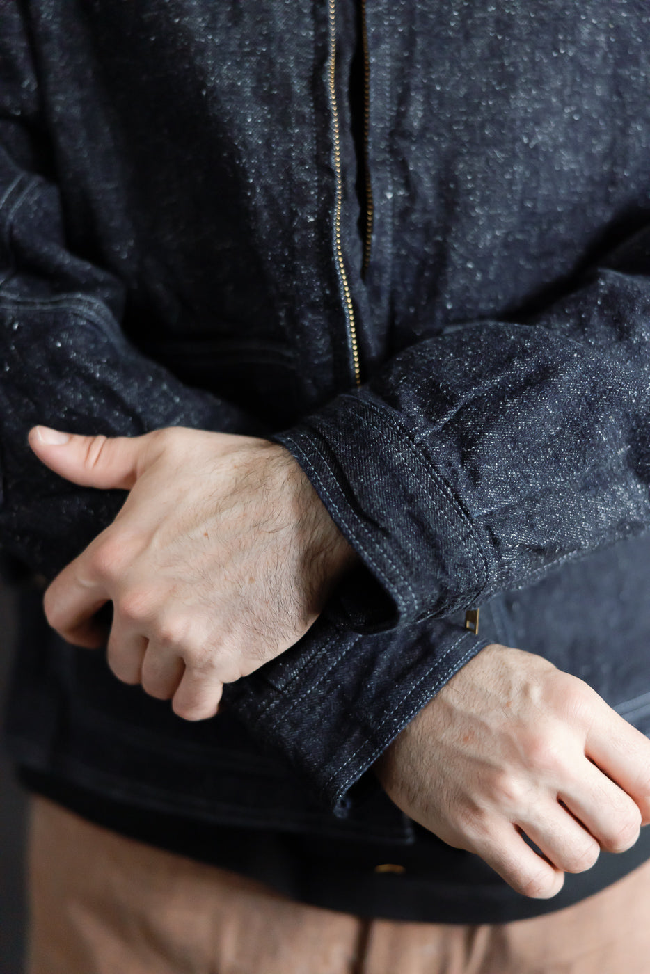 Close-up front view of male model’s dark denim jacket sleeve, showing ribbed cuff, gold zipper, and fabric texture
