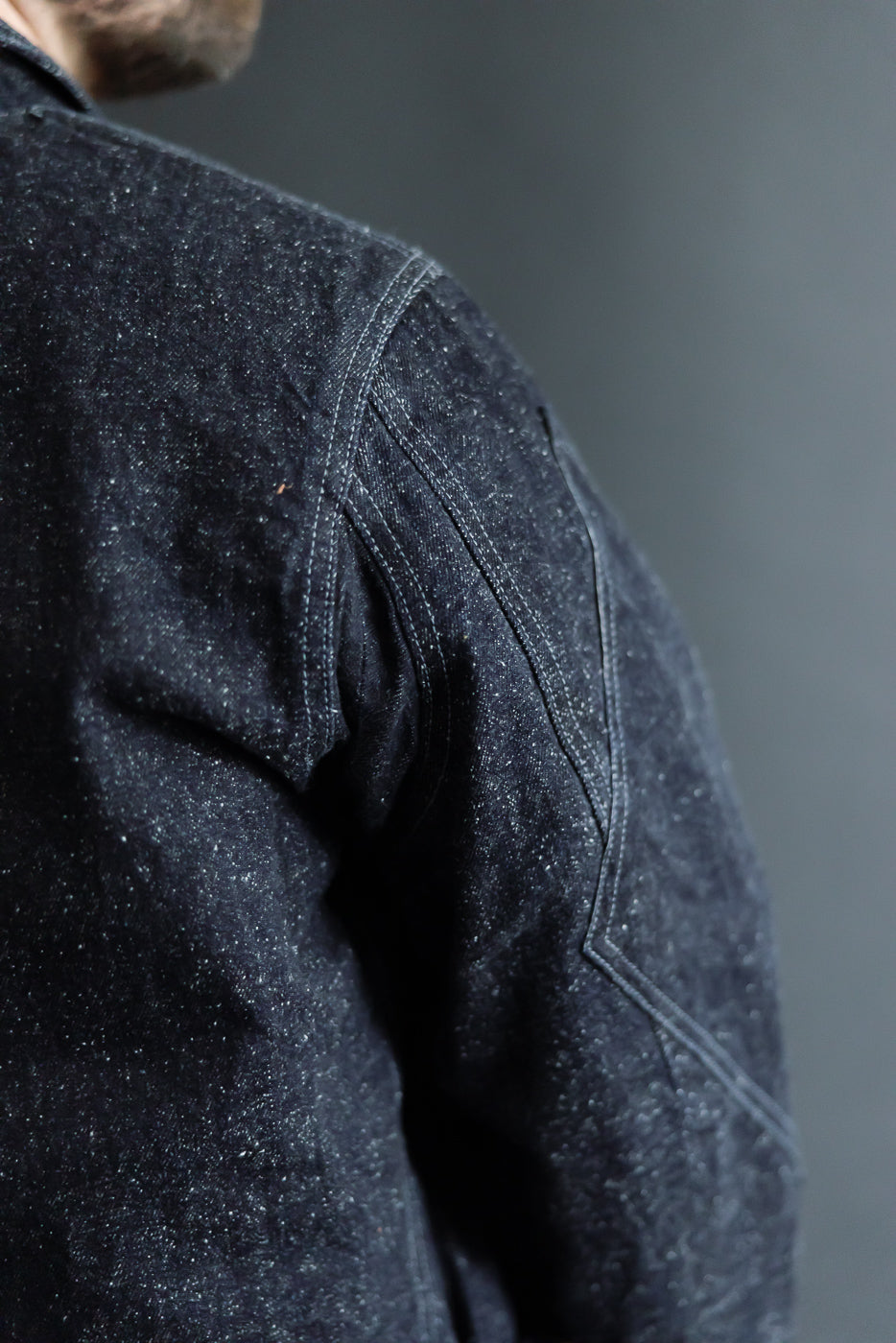 Close-up side view of dark selvedge denim jacket on male model, showing shoulder seam & fabric texture