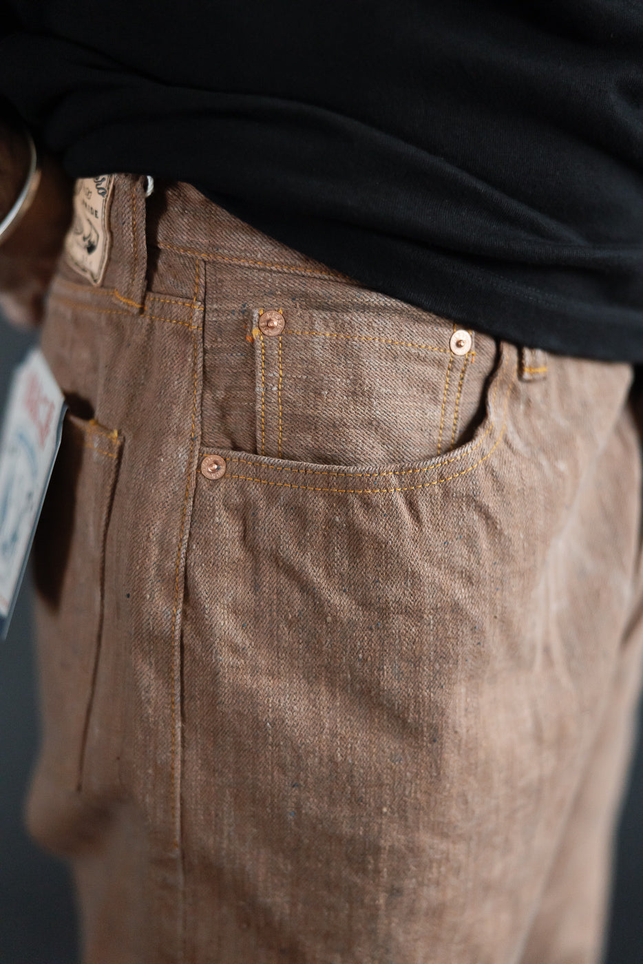 Close-up front view of selvedge denim pants on male model, showing copper rivets, orange stitching, and textured fabric