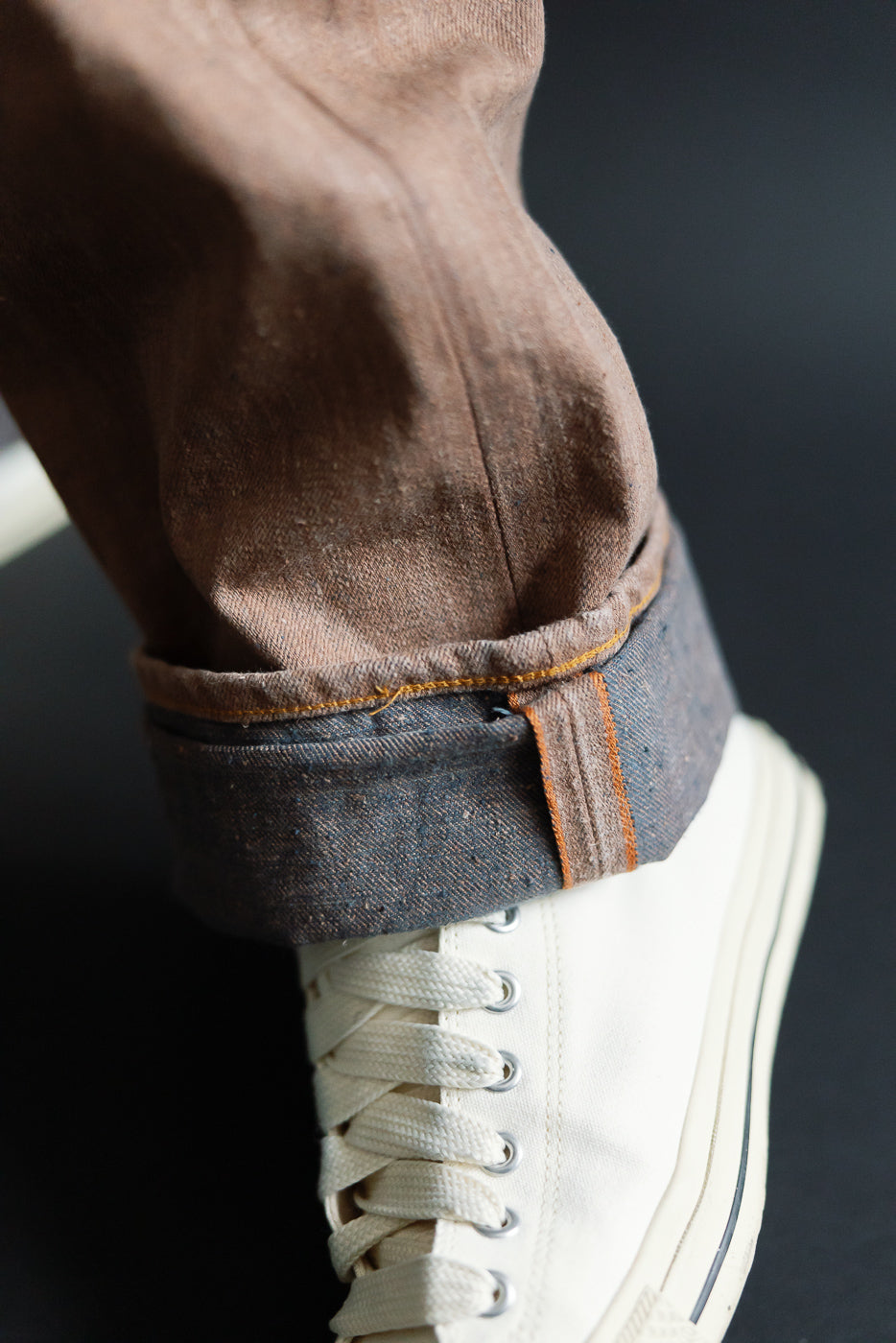 Close-up of male model’s rolled selvedge denim pant cuff, showing orange stitching and fabric texture with white sneaker
