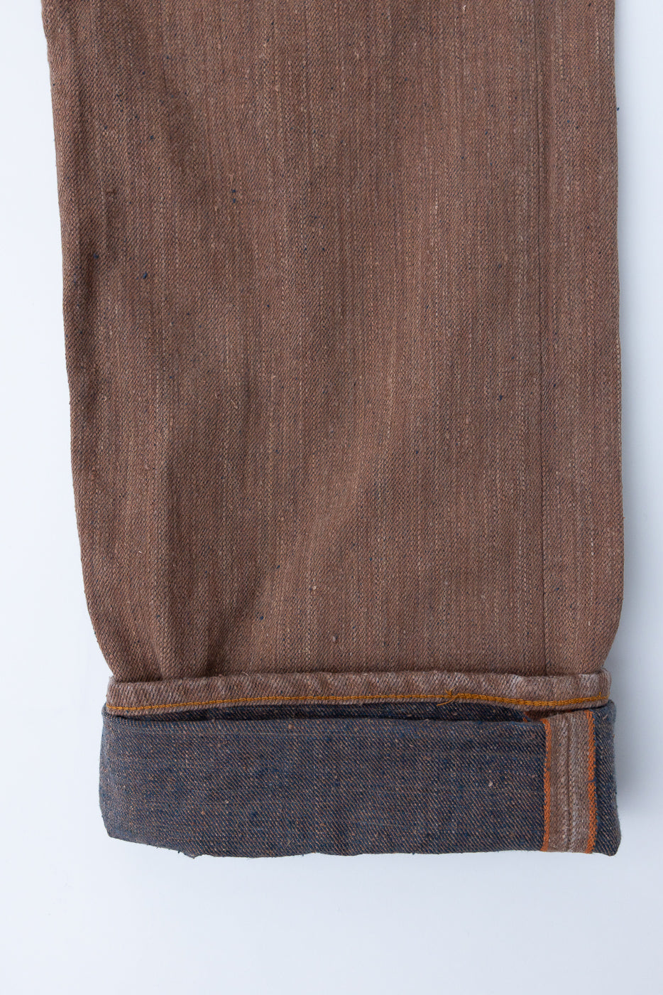 Close-up front view of brown selvedge denim pant leg with rolled cuff, showing texture and orange stitching detail