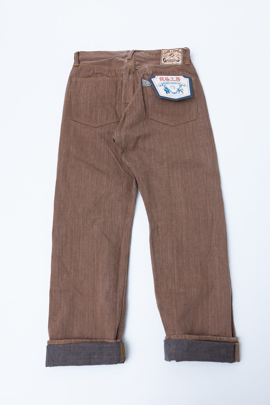 Back view flat lay of brown selvedge denim pants with cuffed hems, back pocket tag, and waist label detail
