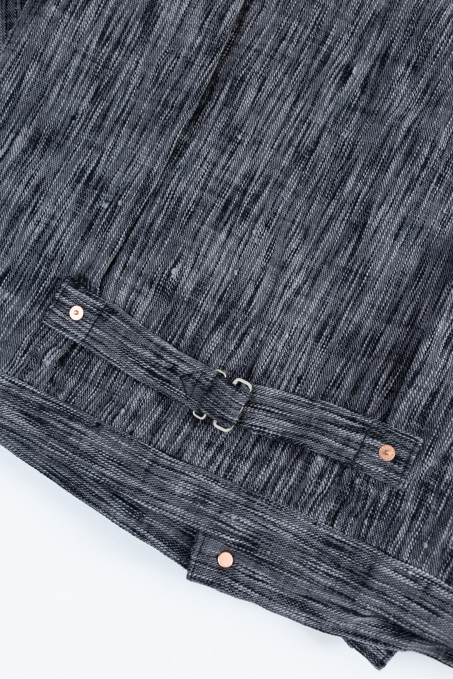 Close-up of dark selvedge denim jacket back, showing textured fabric, copper rivets, and adjustable waist strap detail.