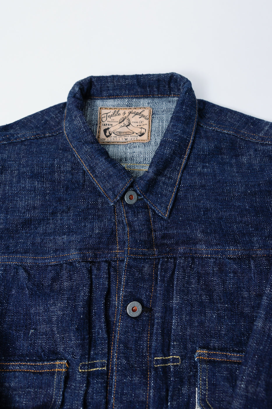 Close-up front view of Graph Zero 15oz indigo slubby selvedge denim jacket collar, buttons, pockets, and leather patch at neckline, highlighting textured fabric and contrast stitching.