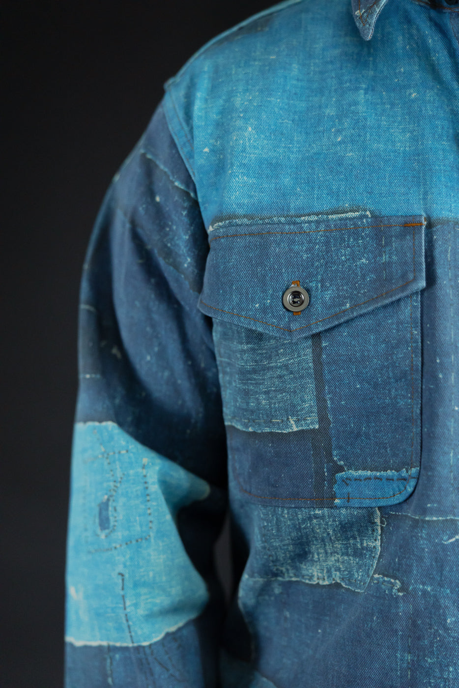 Close-up of male model's left side showing denim shirt pocket with button, patchwork texture, and stitching details.