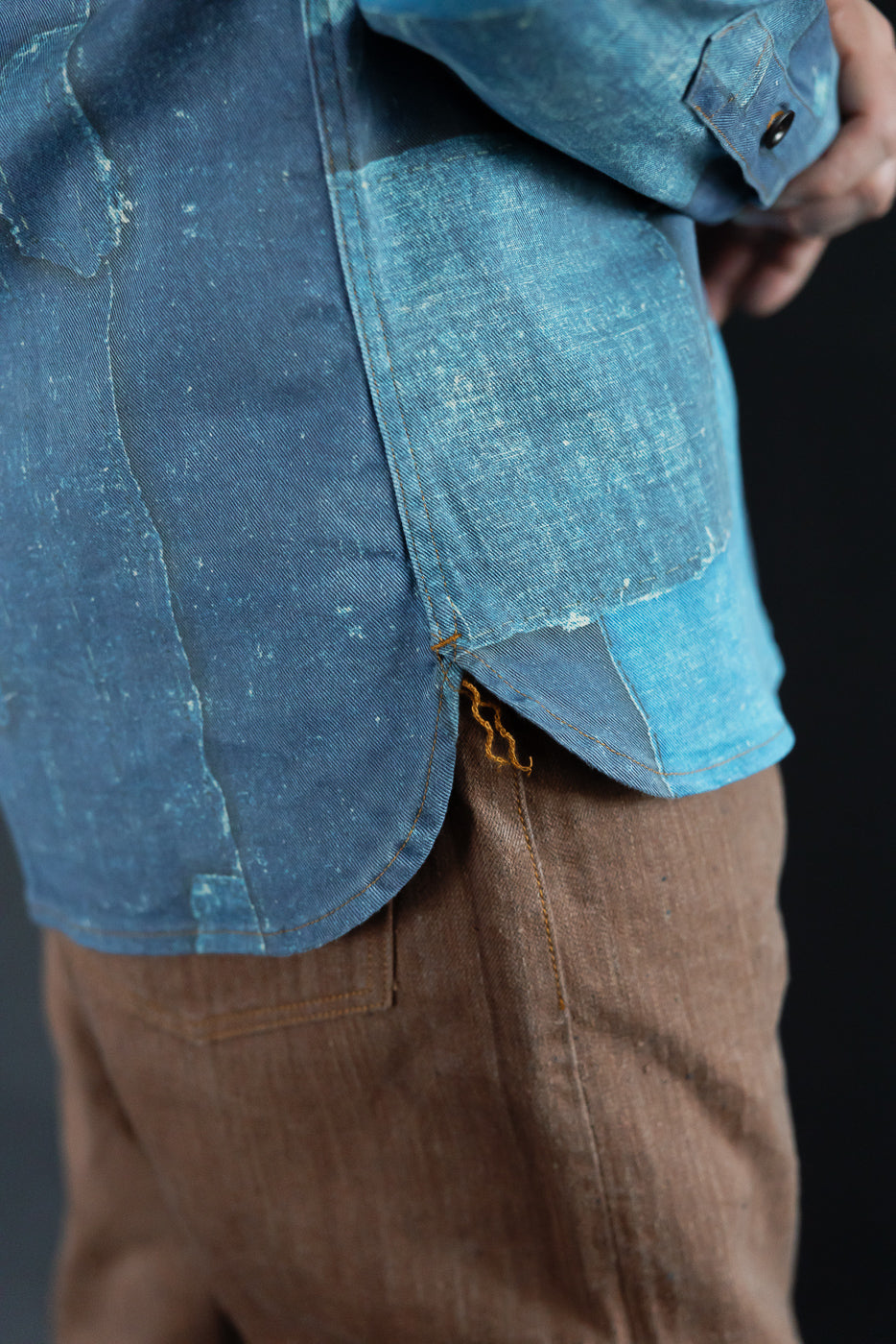 Close-up of male model's side, showcasing denim shirt hem with orange stitching and patchwork detail.