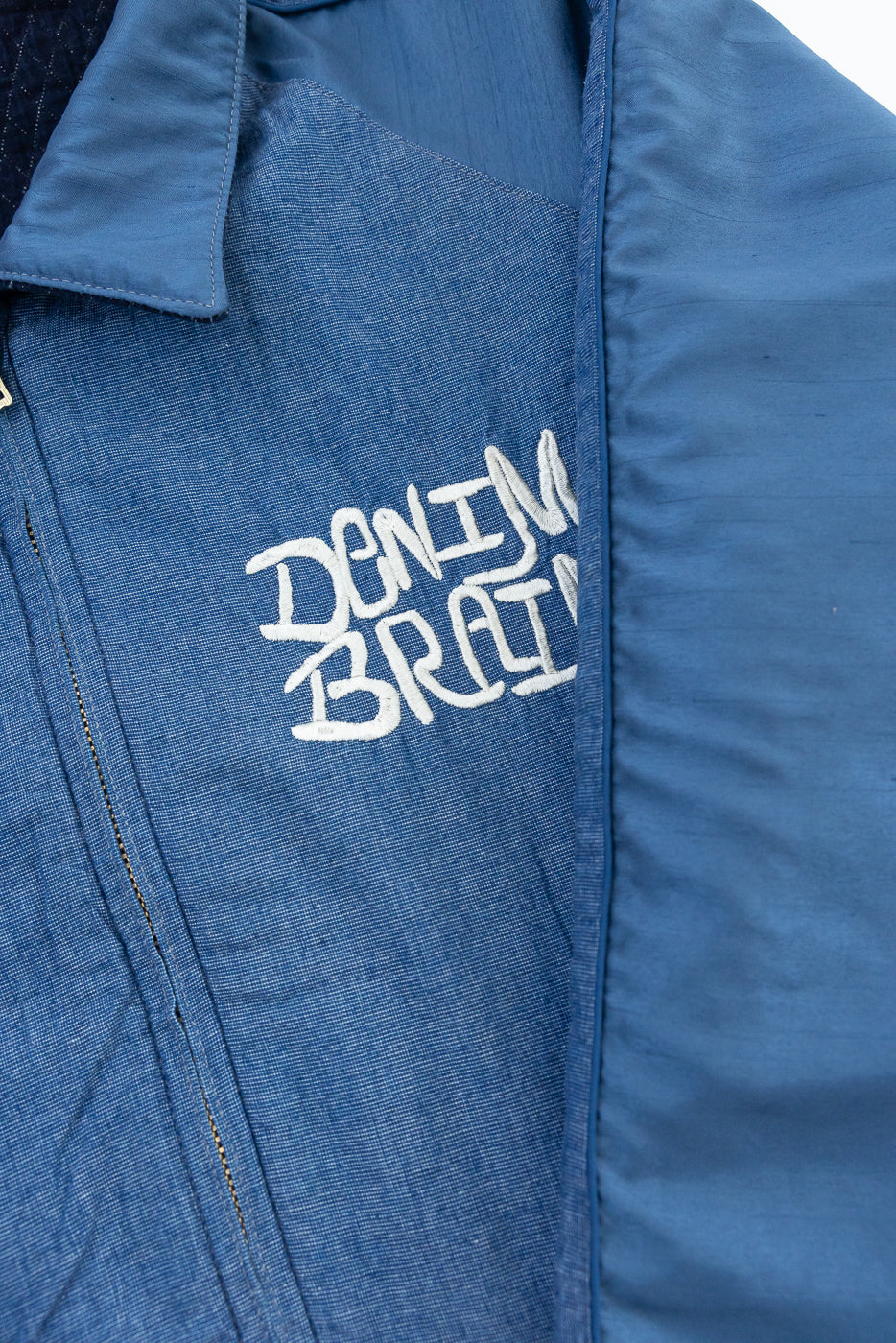 Close-up of blue denim jacket front, showing 'DENIM BRAIN' embroidery, collar, and fabric texture.
