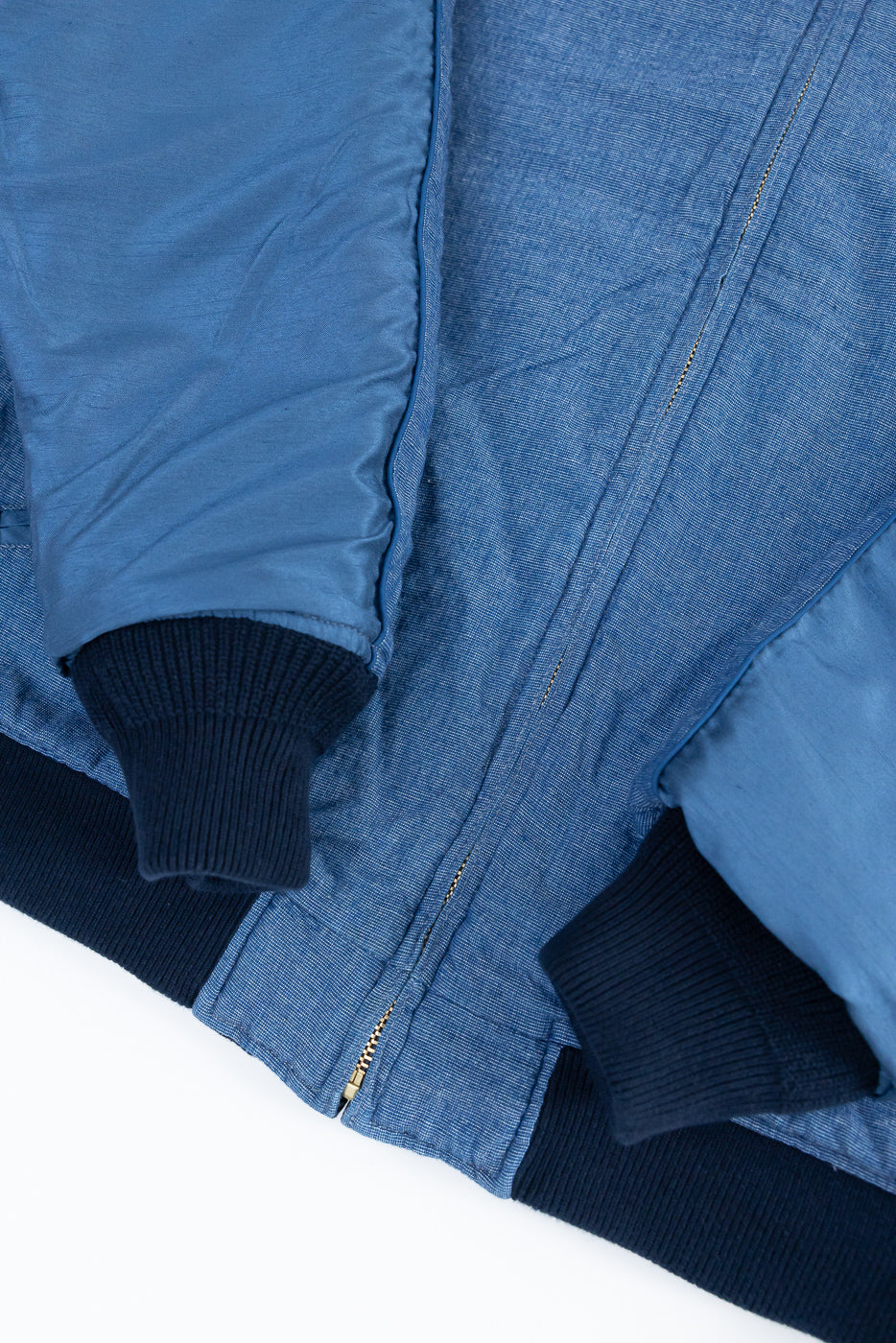 Close-up front view of blue denim bomber jacket with ribbed cuffs, gold zipper, and contrast stitching