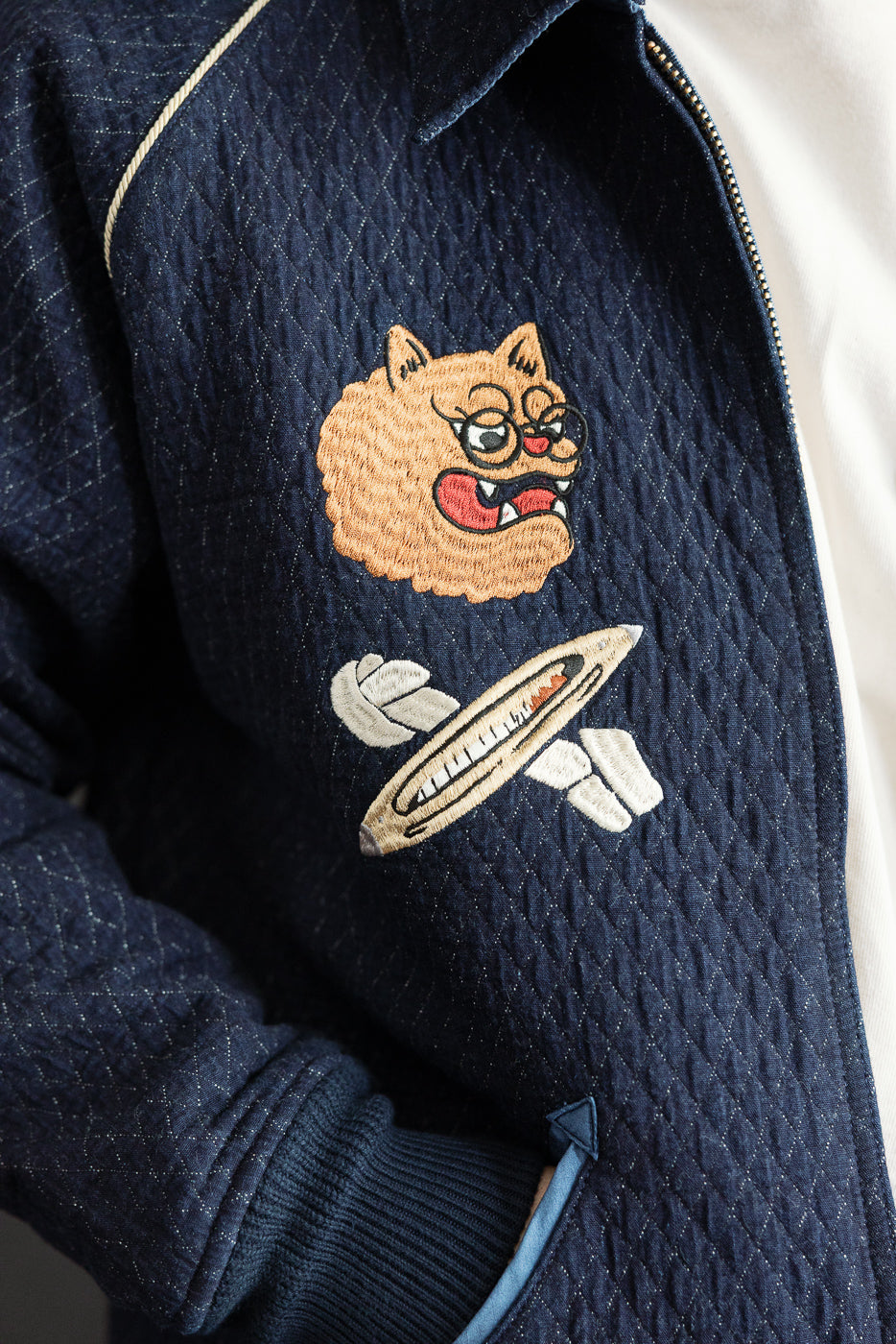 Close-up front view of navy quilted jacket with embroidered cat and surfboard patch, ribbed cuffs, zipper detail
