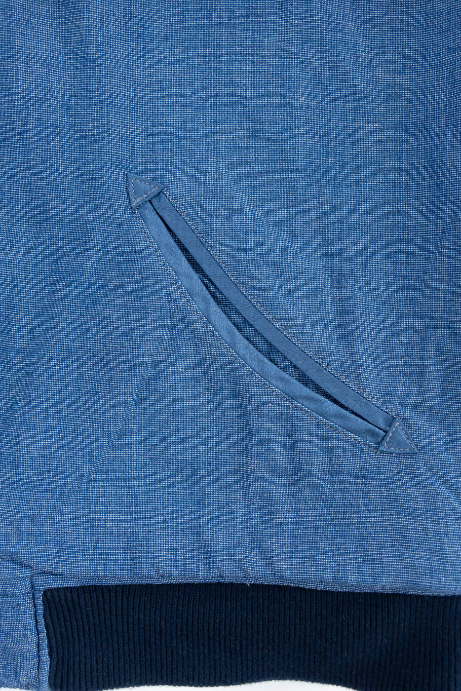 Close-up of blue chambray shirt front, showing diagonal chest pocket with stitched triangle detail and ribbed hem.