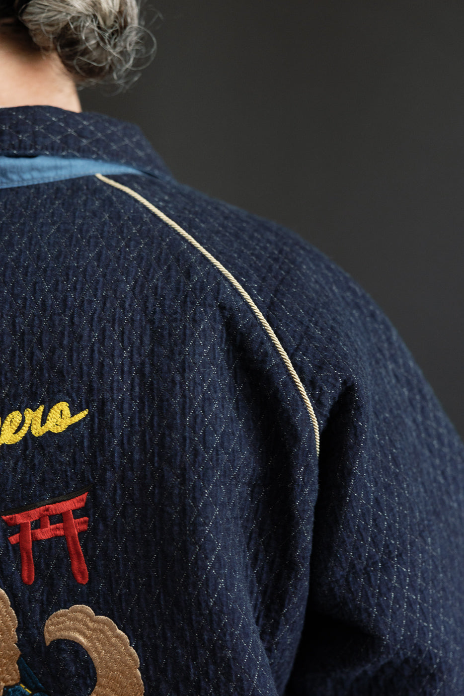 Close-up back view of men's indigo quilted jacket with embroidery, gold piping, and textured fabric.