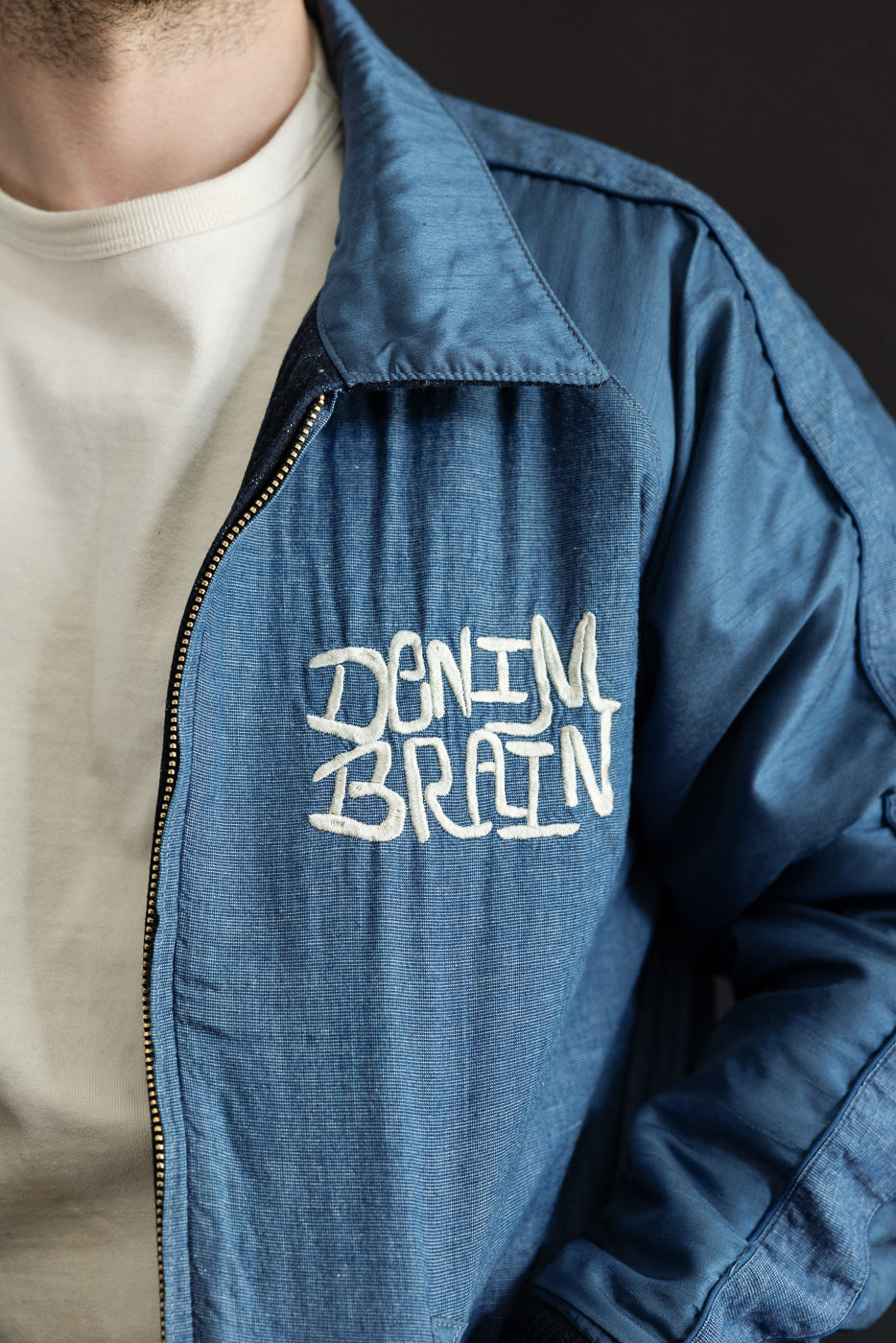 Close-up front view of male model wearing blue denim jacket with 'DENIM BRAIN' embroidery, showing collar and zipper detail.