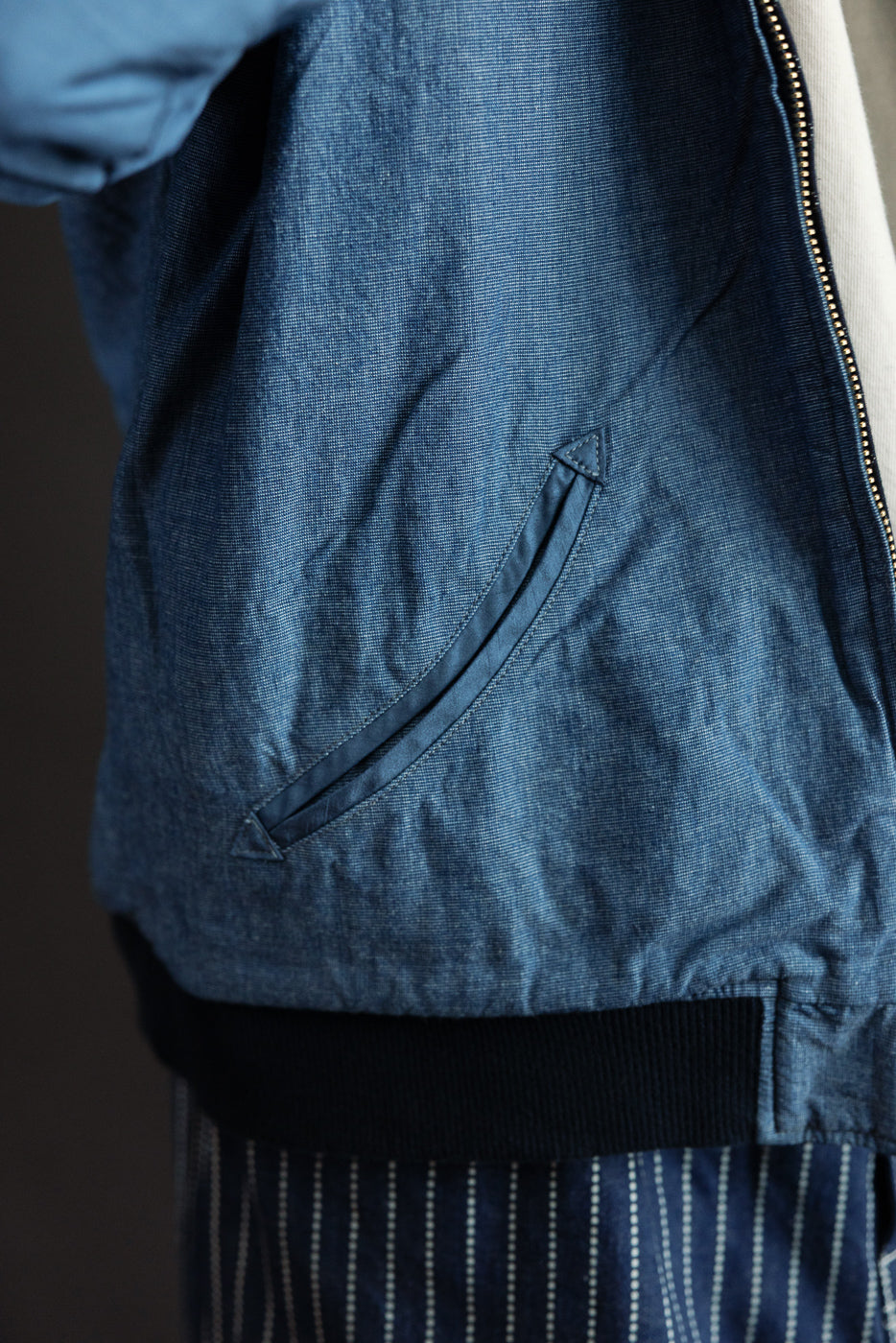 Close-up side view of men's denim bomber jacket showing diagonal chest pocket, fabric texture, and ribbed hem
