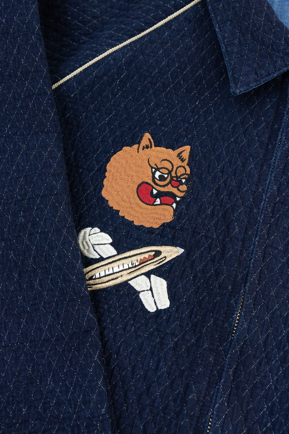 Close-up front of navy quilted jacket with embroidered cat graphic, diamond stitch texture, and contrast piping