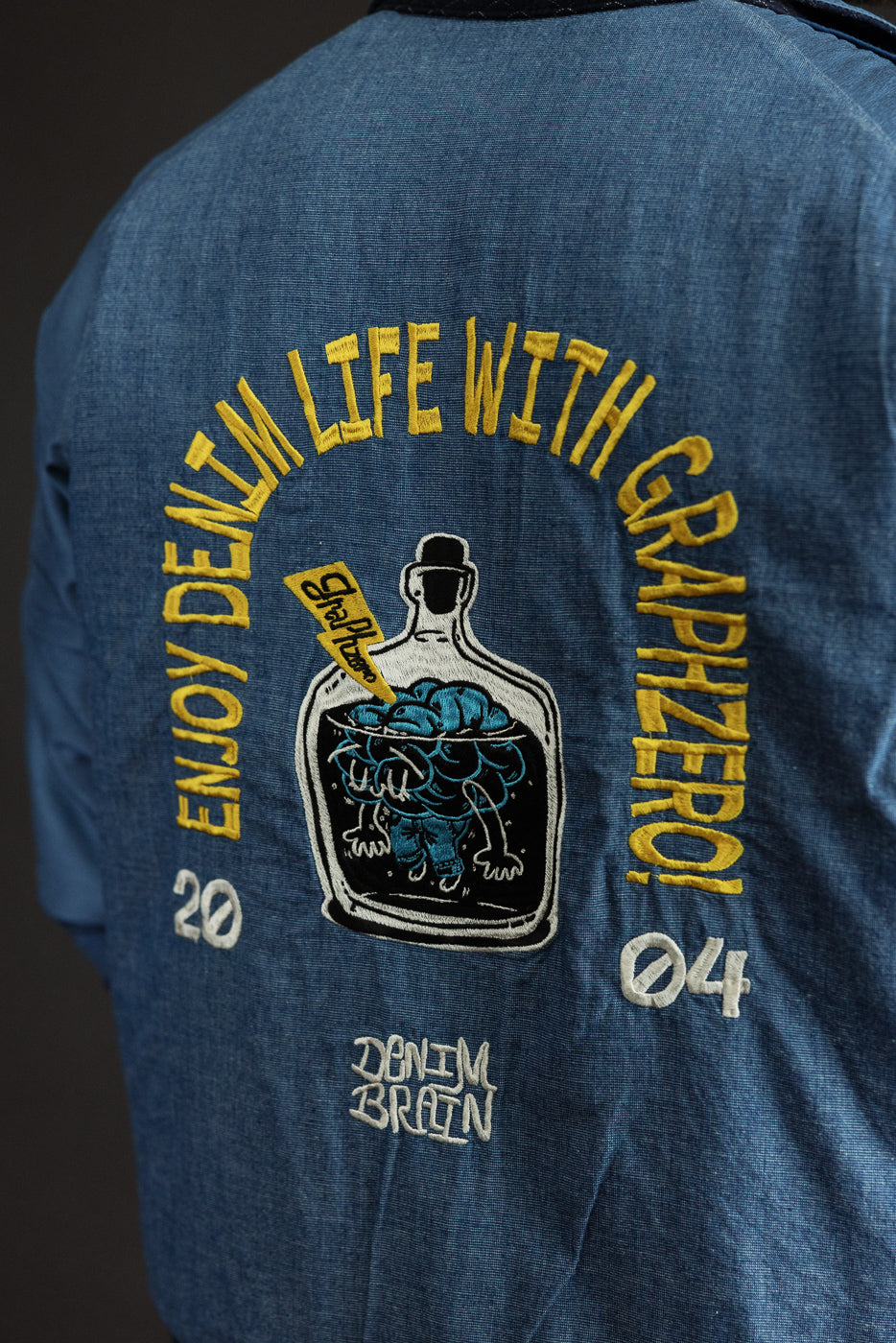 Back view of denim jacket with embroidered 'Enjoy Denim Life' graphic, featuring bottle design and 'Denim Brain' text.