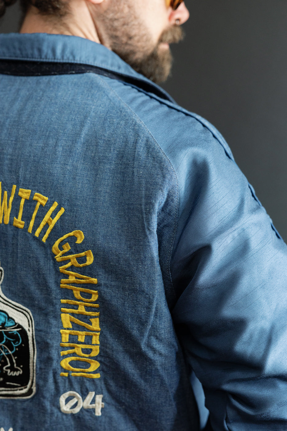 Close-up back view of male model in blue denim jacket with yellow 'WITH GRAPHZERO!' embroidery.