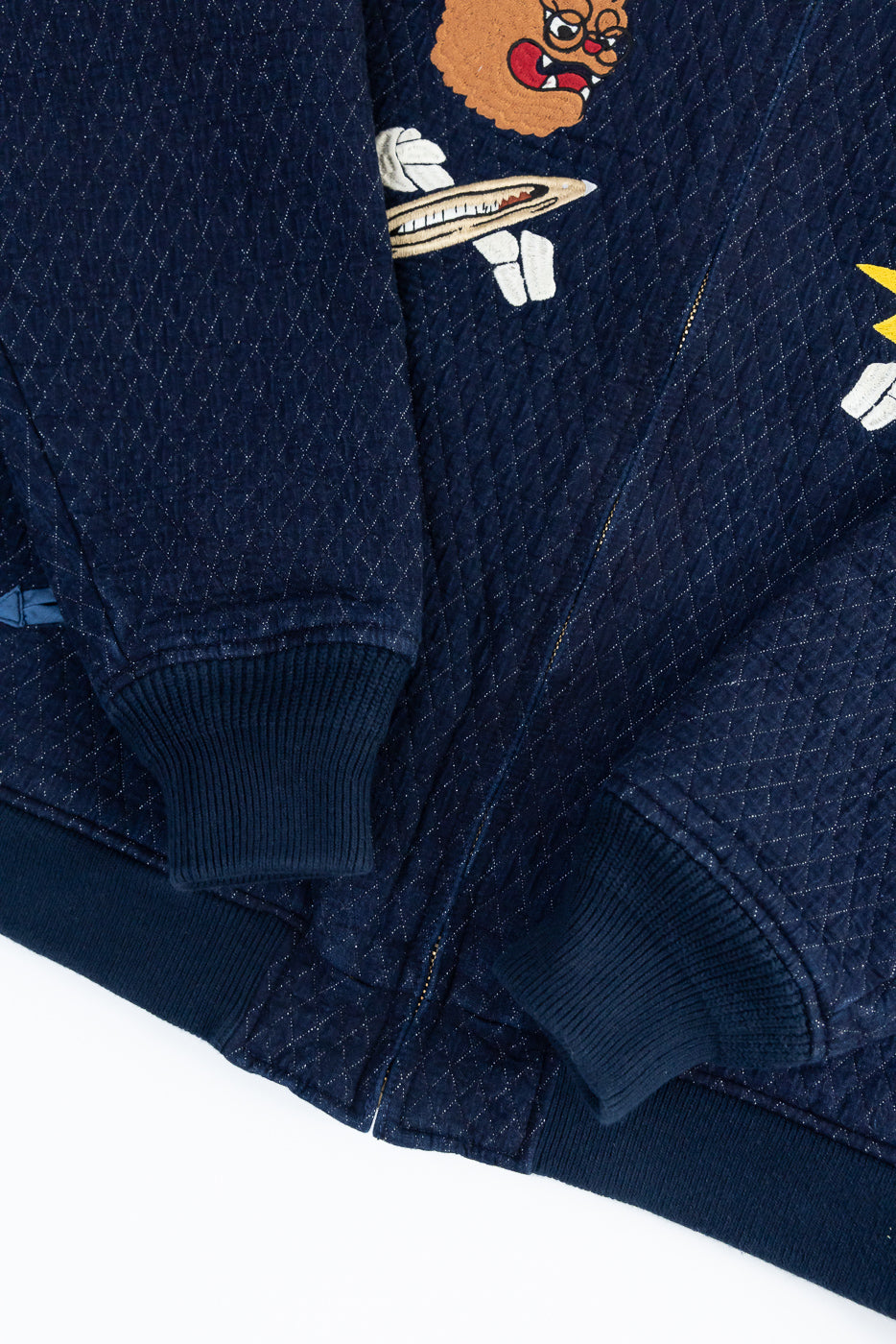 Close-up of navy quilted jacket front, showcasing embroidered design, ribbed cuffs, and diamond stitching texture.