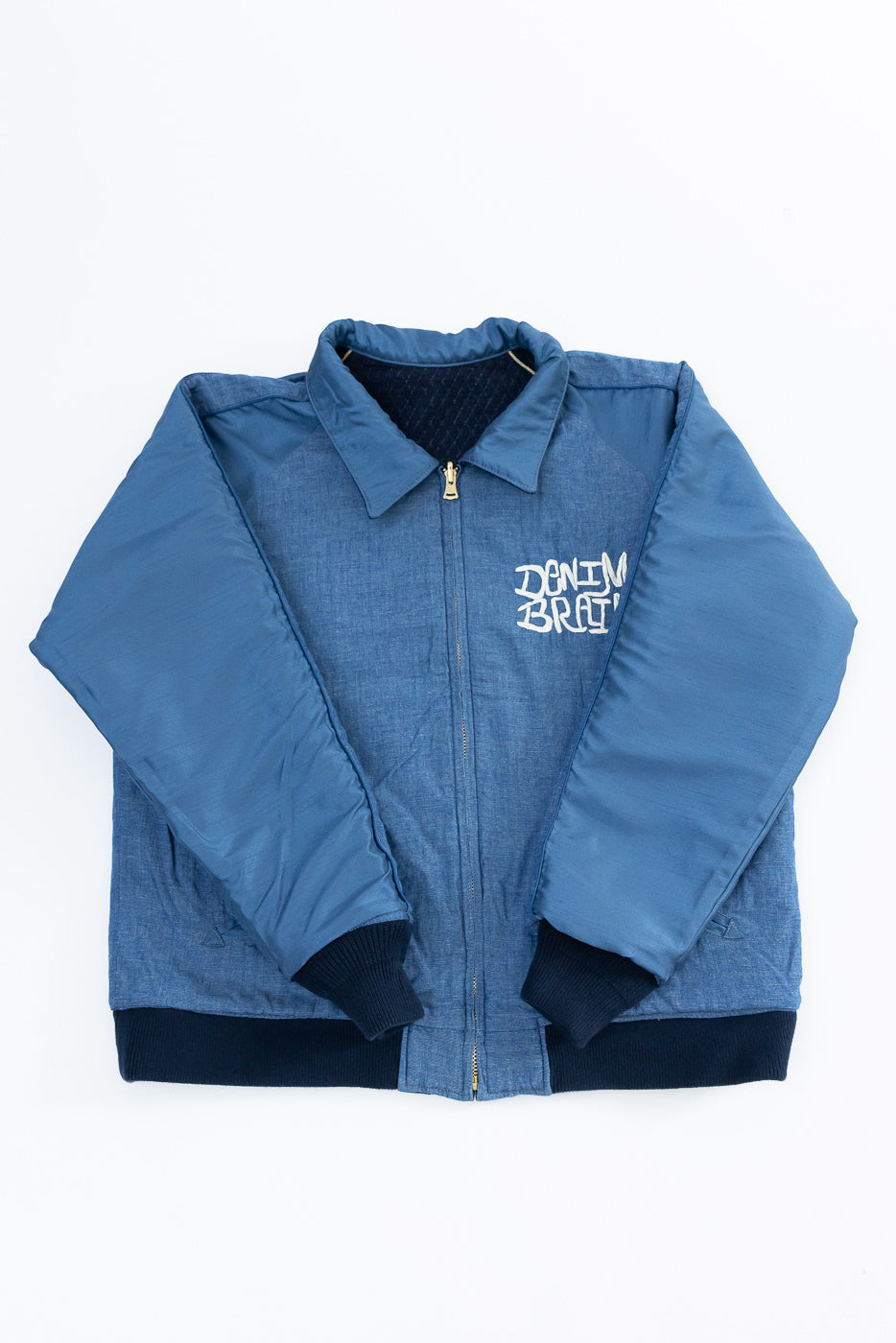 Flat lay front view of blue denim bomber jacket with 'DENIM BRAT' graphic, ribbed cuffs, and collar