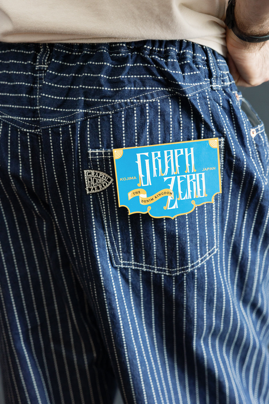 Close-up of back pocket on striped denim pants, showing Graph Zero label, worn by male model.