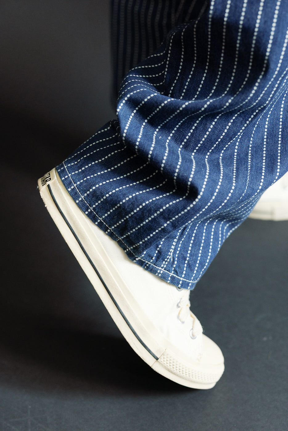 Close-up of male model's lower leg in navy pinstripe selvedge jeans, showing hem and white sneaker