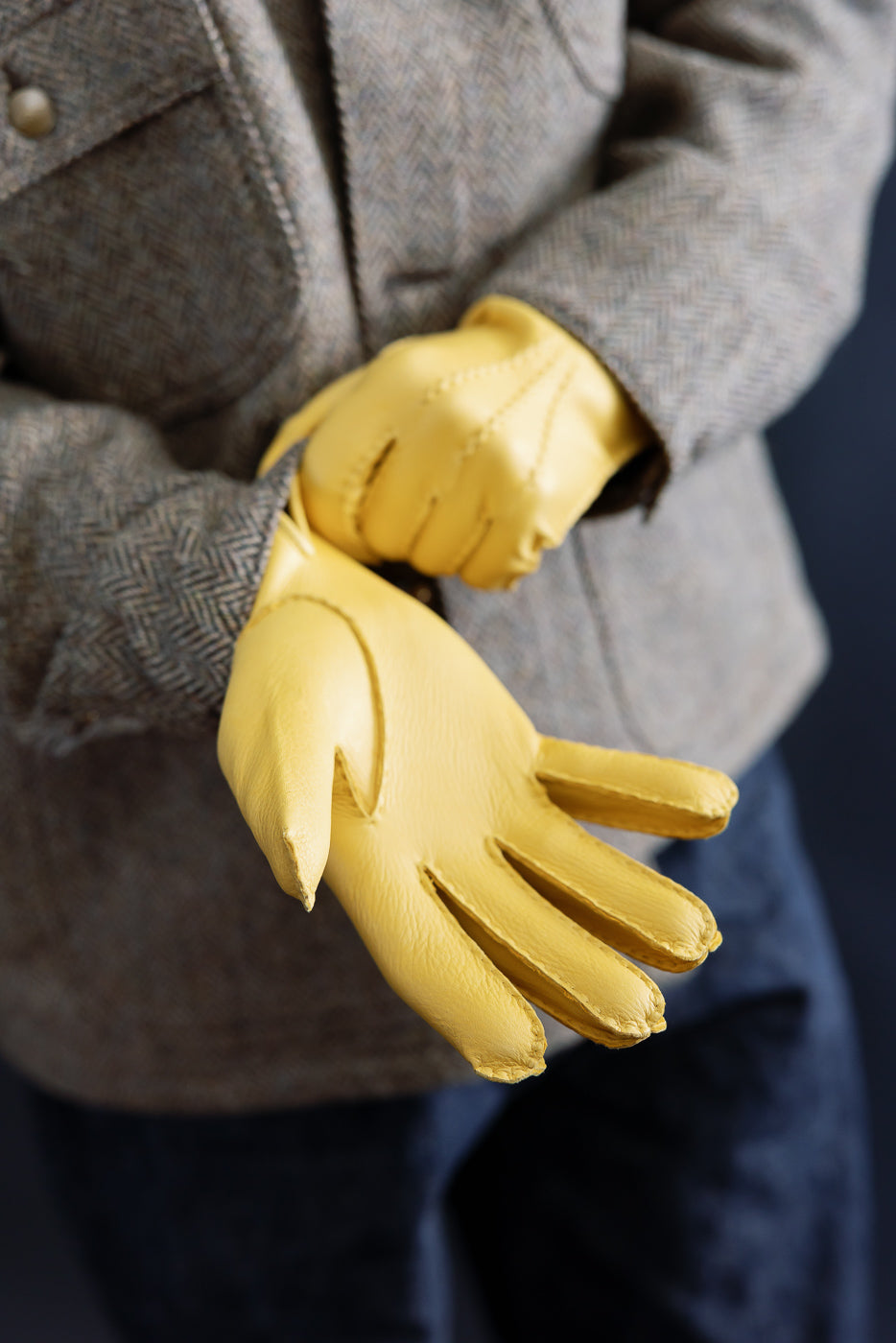 Close-up of men's Hestra Henry natural yellow leather gloves on model, front hand view, textured jacket visible, shot highlights glove stitching and leather grain.
