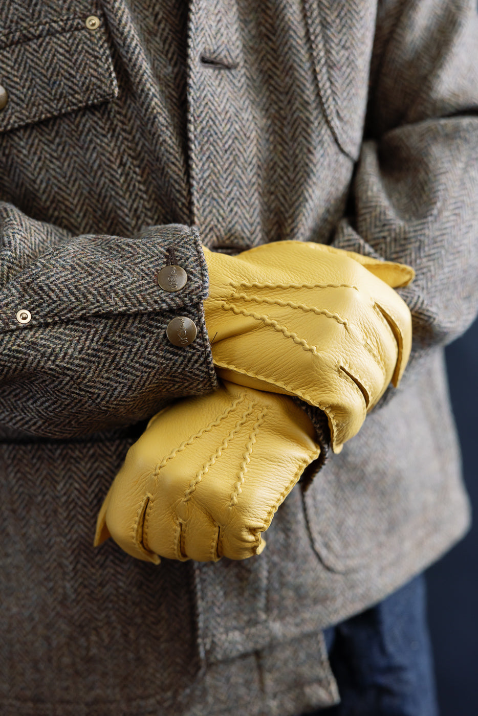 Close-up of Hestra Henry natural yellow leather gloves, front view, worn by male model wearing herringbone jacket, showcasing glove texture and stitching detail.