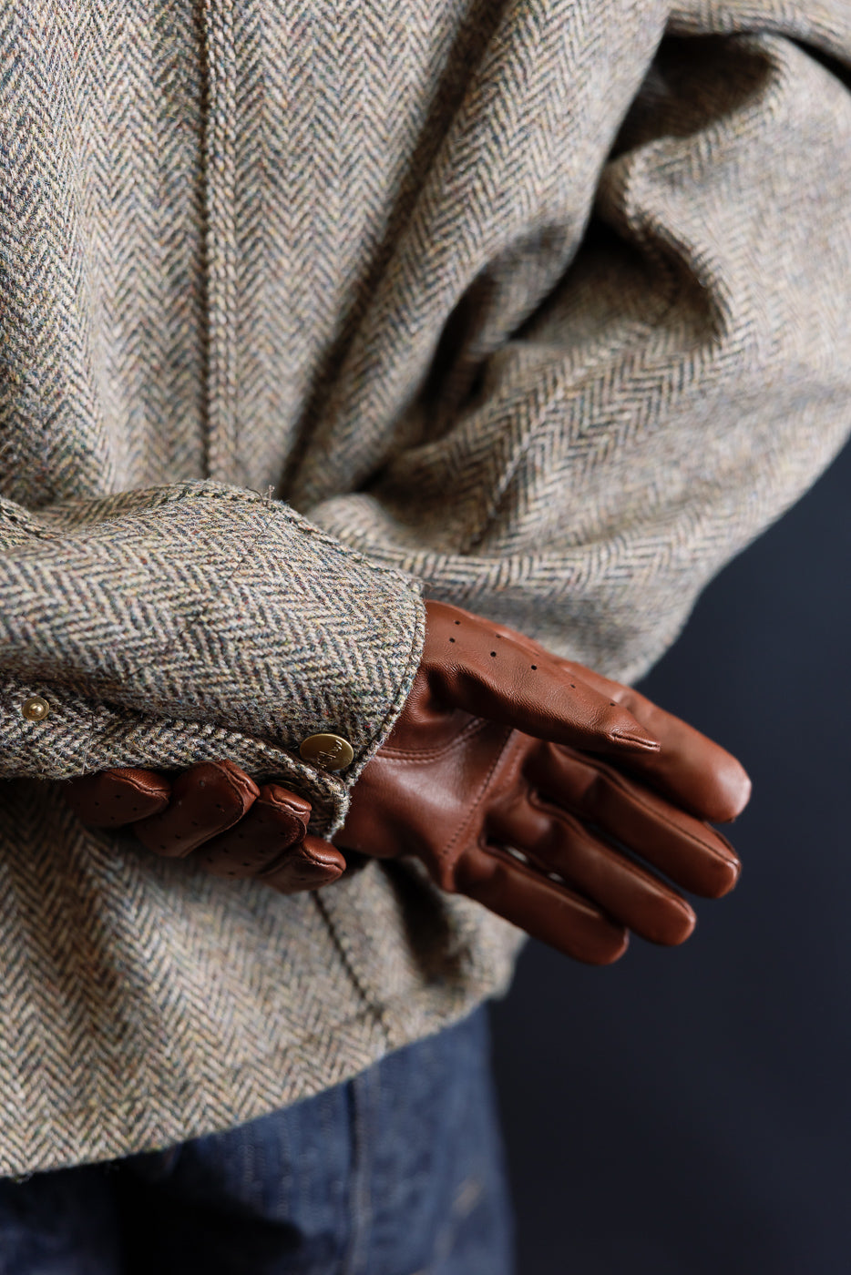 Close-up of chestnut brown Hestra Morgan driving glove on male model’s right hand, showing glove back and jacket sleeve details.