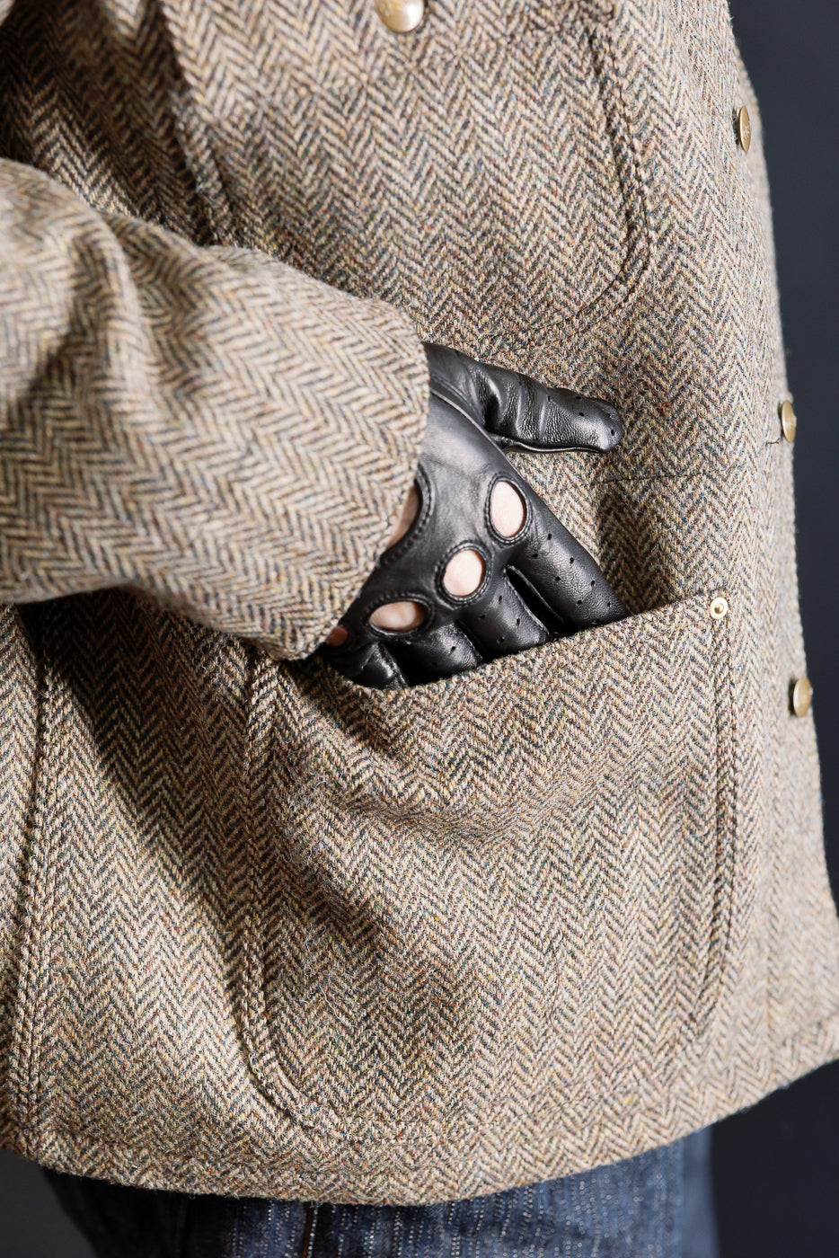 Close-up of male model’s right side herringbone tweed jacket pocket, black leather driving glove, gold snap buttons, Dant contemporary casual accessory.