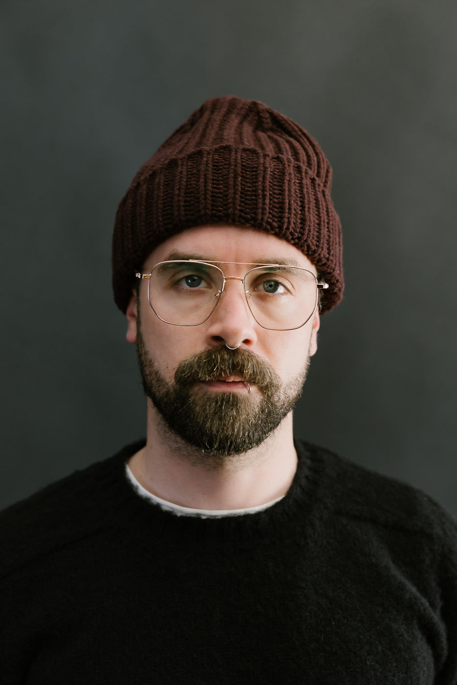 Man wearing a brown knit beanie and glasses against a dark background