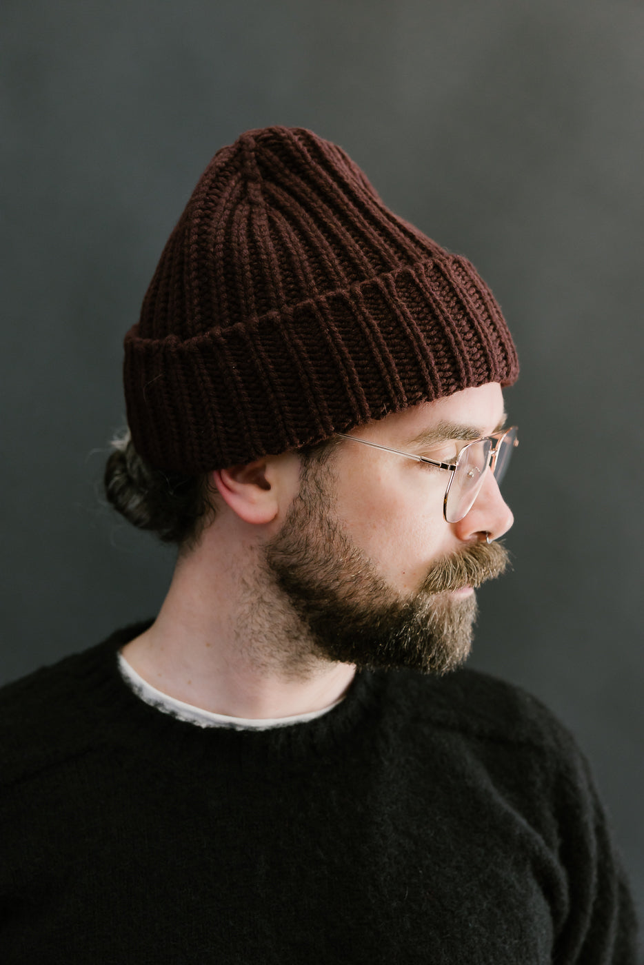 Man wearing a brown knit beanie and glasses against a dark background
