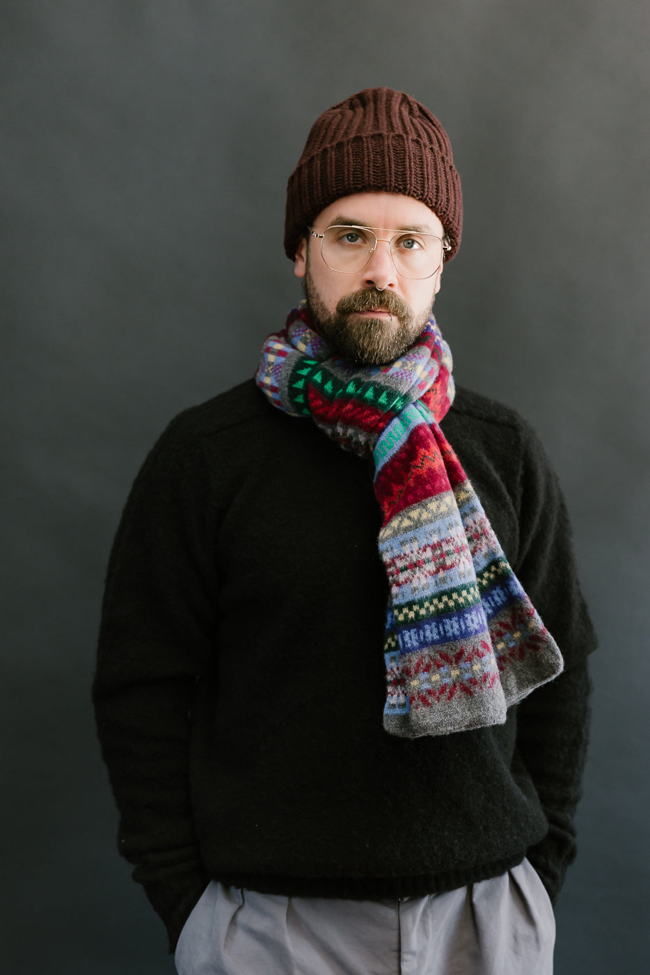multicolored, patterned wool scarf with a heather grey base on a bearded man with a black sweater and chocolate hat, front view