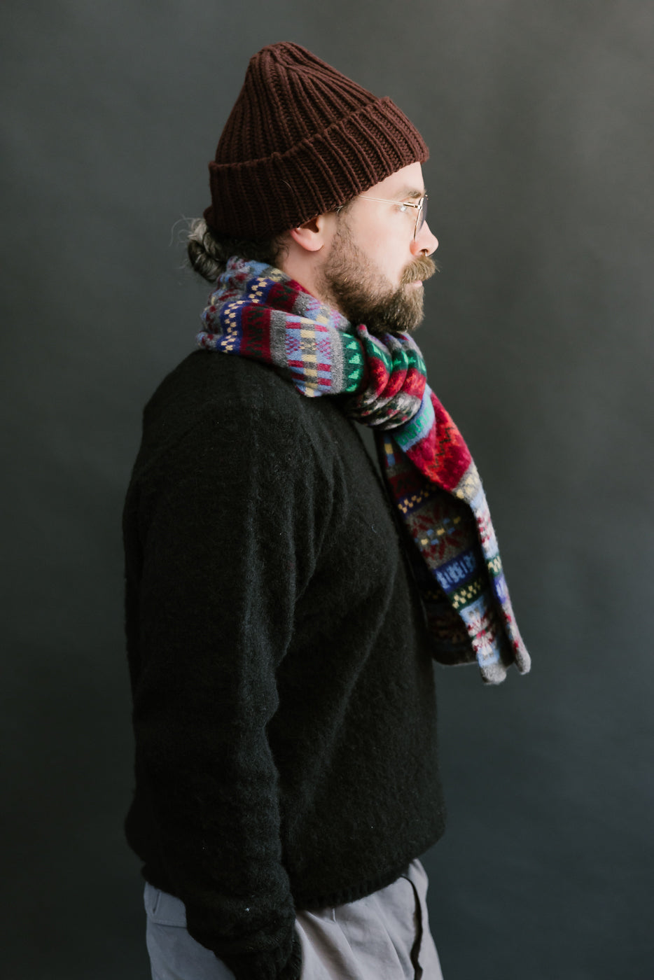 multicolored, patterned wool scarf with a heather grey base on a bearded man with a black sweater and chocolate hat, side view