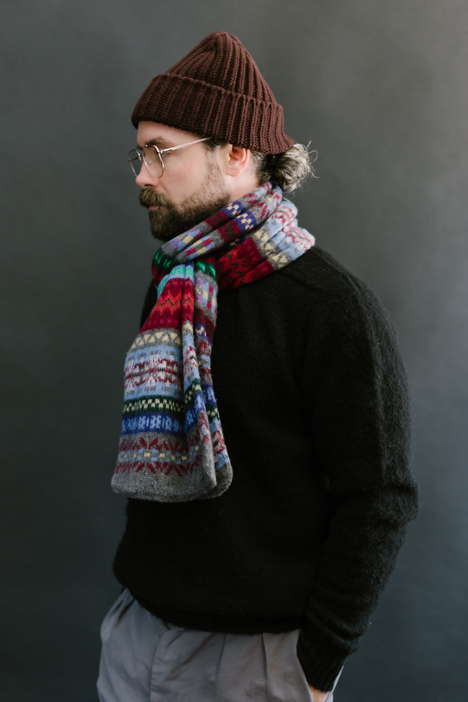 multicolored, patterned wool scarf with a heather grey base on a bearded man with a black sweater and chocolate hat, side view