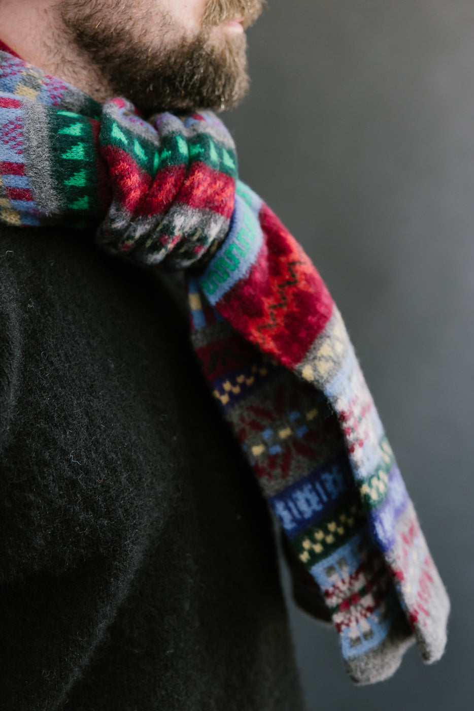 multicolored, patterned wool scarf with a heather grey base on a bearded man with a black sweater and chocolate hat, close up, side view