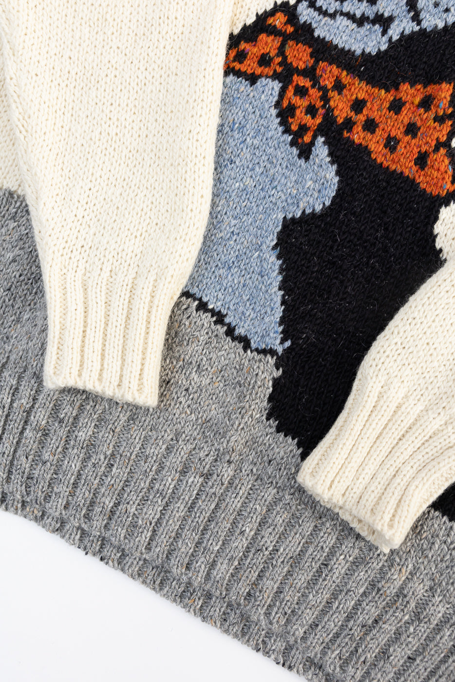 Close-up of front hem and sleeves of knit sweater showing ribbed cuffs, textured wool, and colorful graphic pattern. No model present.