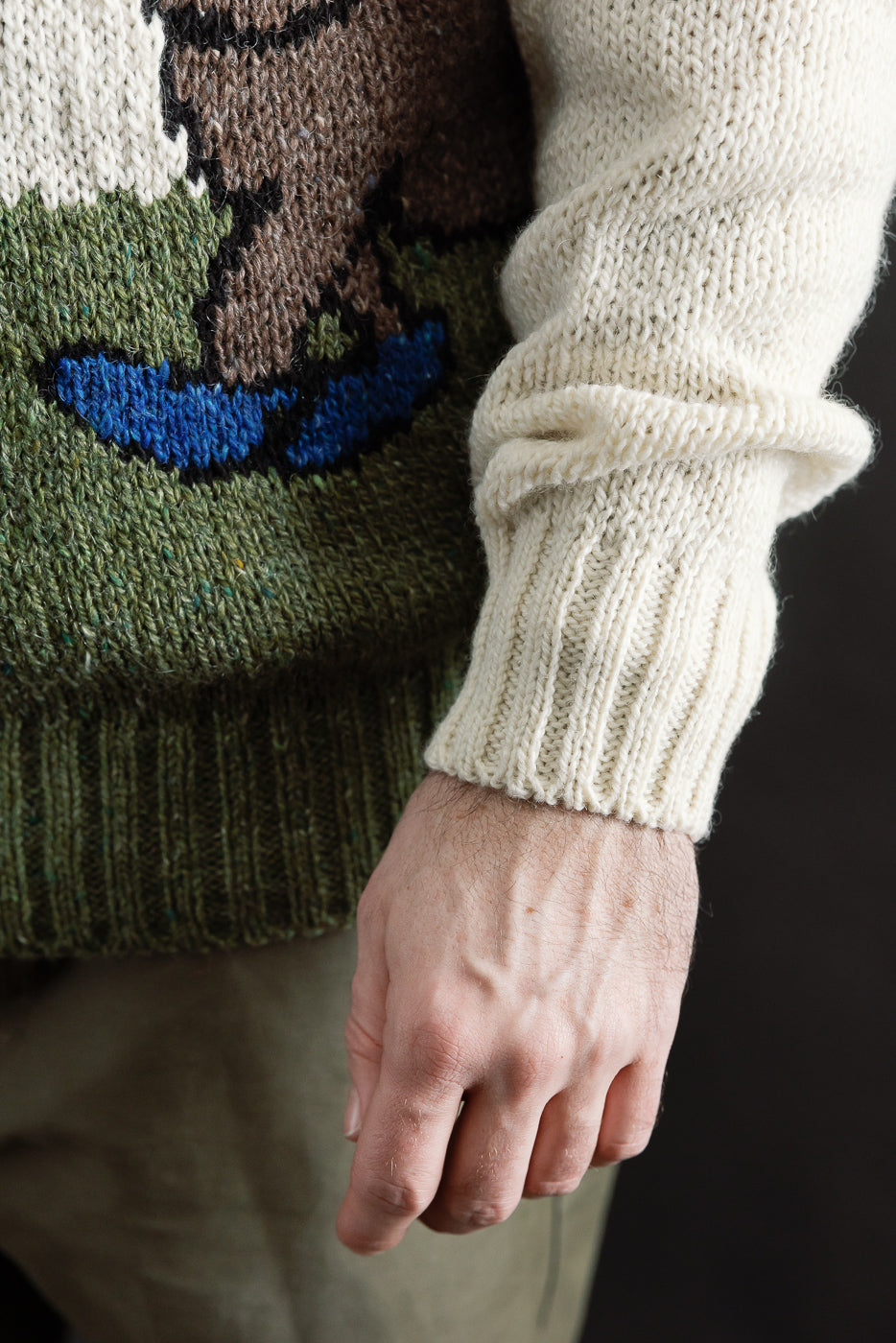 Close-up of male model's right side showing chunky knit sweater sleeve, ribbed cuff, and graphic pattern. Model is 5'9