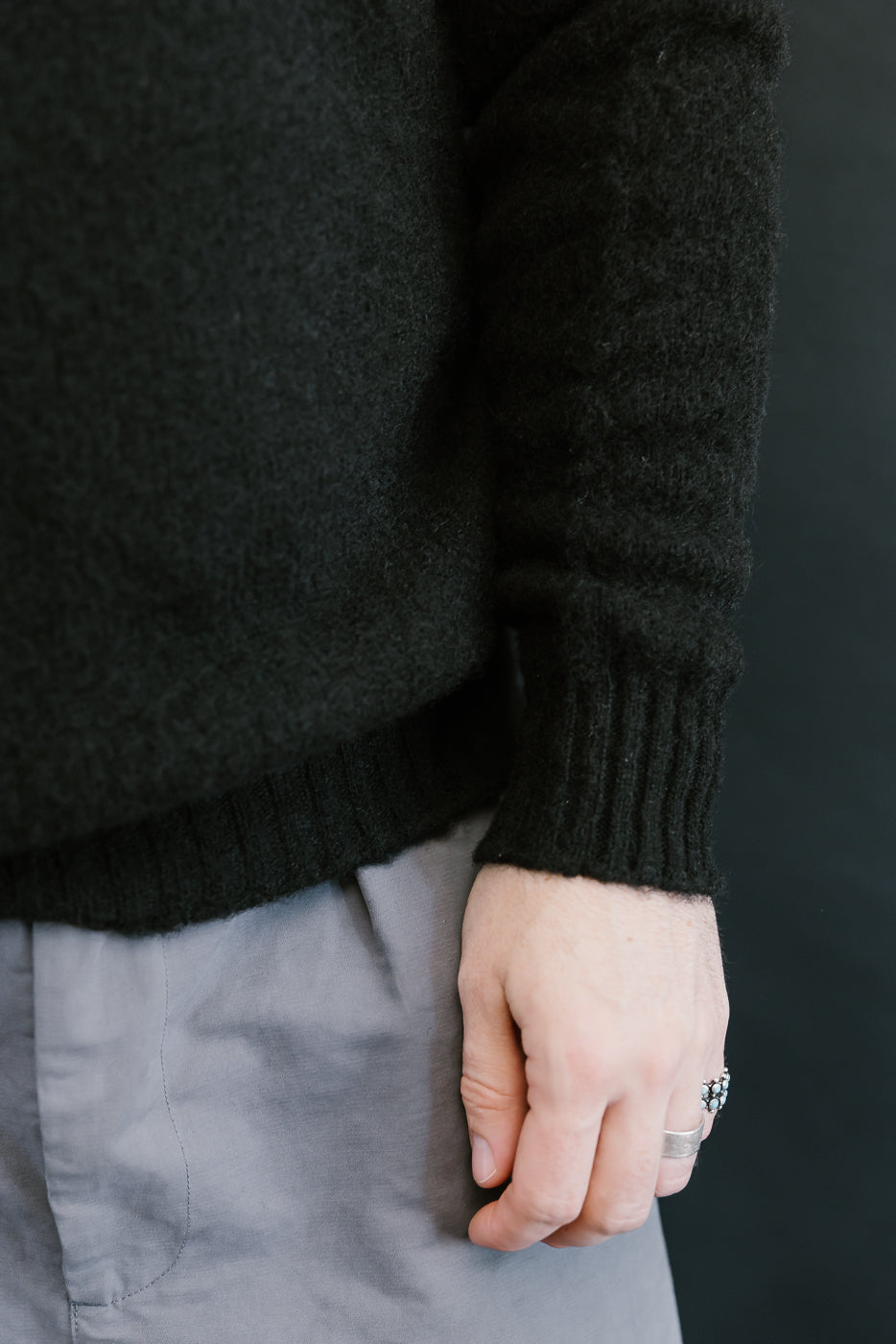 Person wearing a dark sweater and light pants against a black background