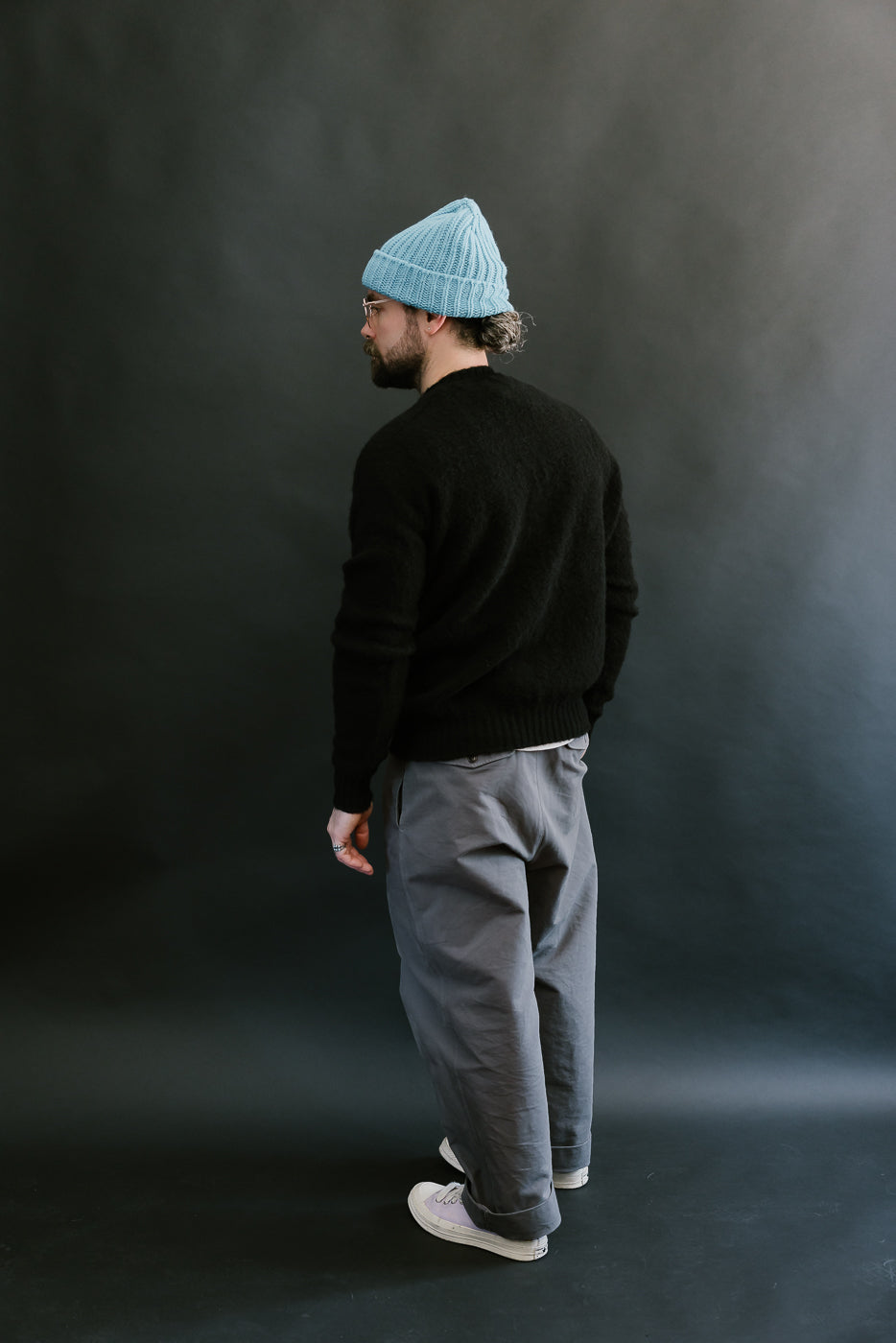 Person wearing a black sweater, gray pants, and a blue knit beanie against a dark background
