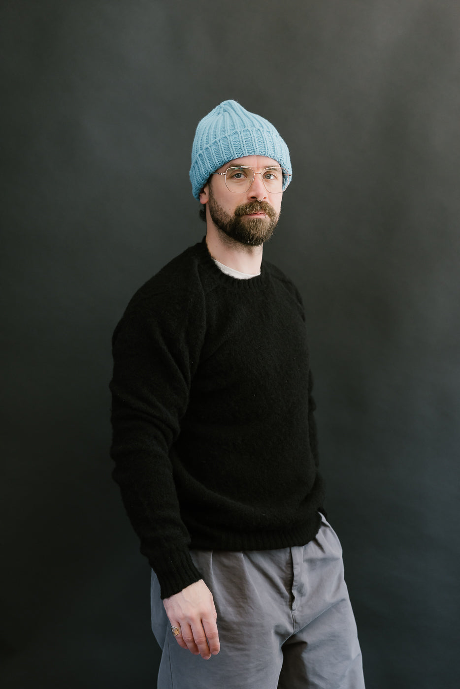 Man wearing a black sweater and gray pants with a blue knit beanie against a dark background