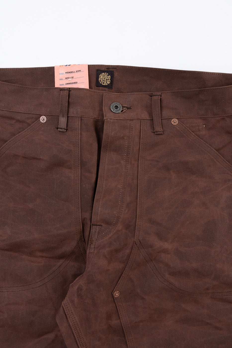 Close-up front view of the waistband and fly of brown unwashed selvedge denim pants, showing the button, belt loops, and a pink tag.