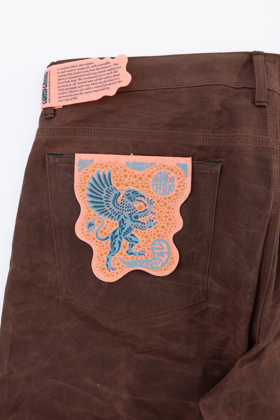 Close-up of the back right pocket of brown pants featuring a blue griffin patch and tag.
