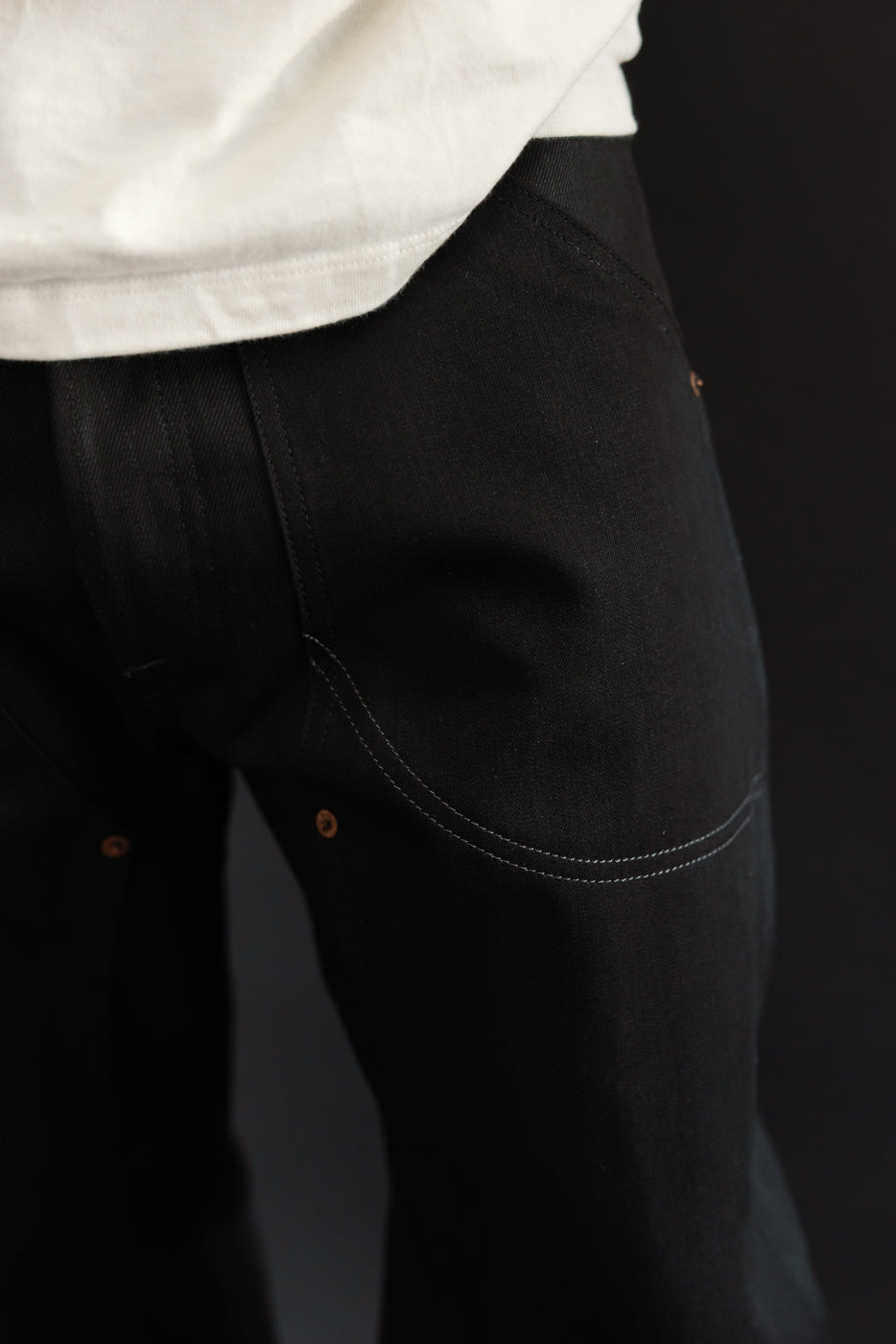 Close-up of black selvedge jeans' back pocket, showing white stitching and copper rivets, worn by male model.
