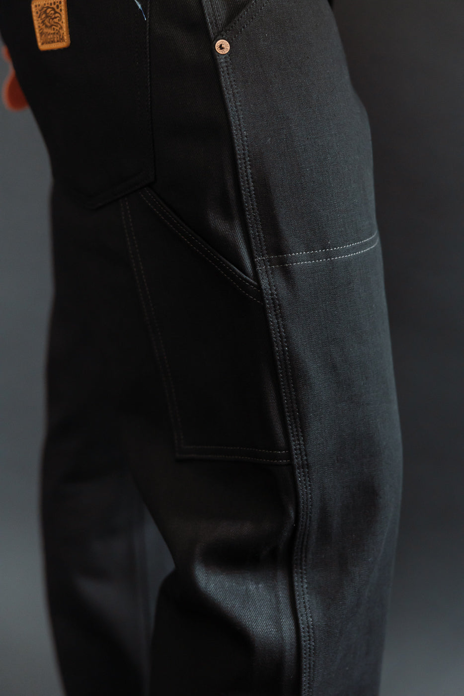 Close-up side view of black selvedge work pants, showing pocket, stitching, and tag detail on male model.