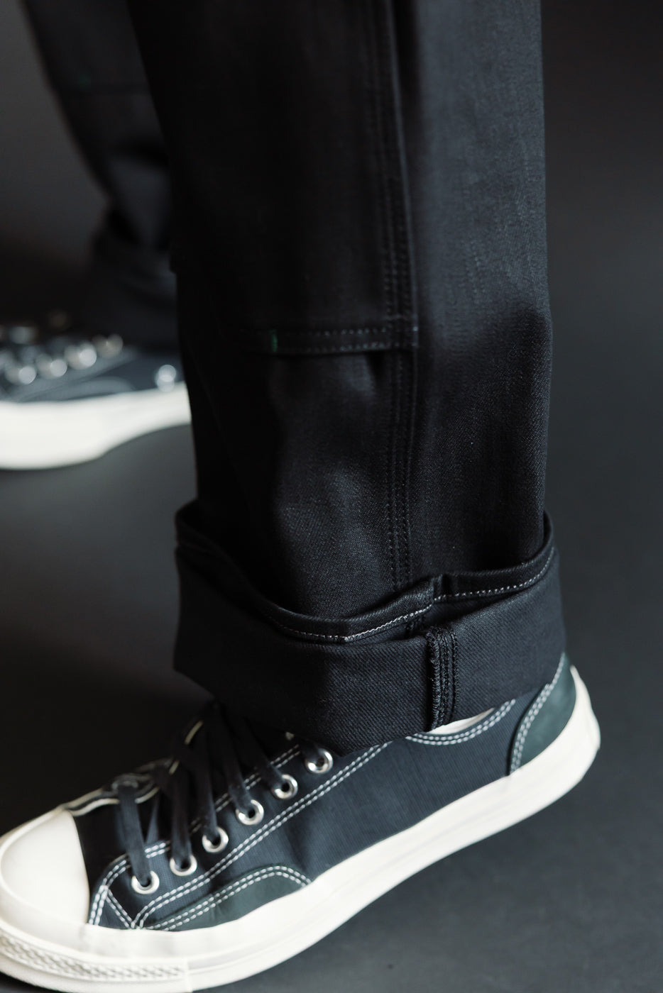 Close-up of male model's lower leg showing black selvedge jeans with cuffed hem and white-stitched sneakers
