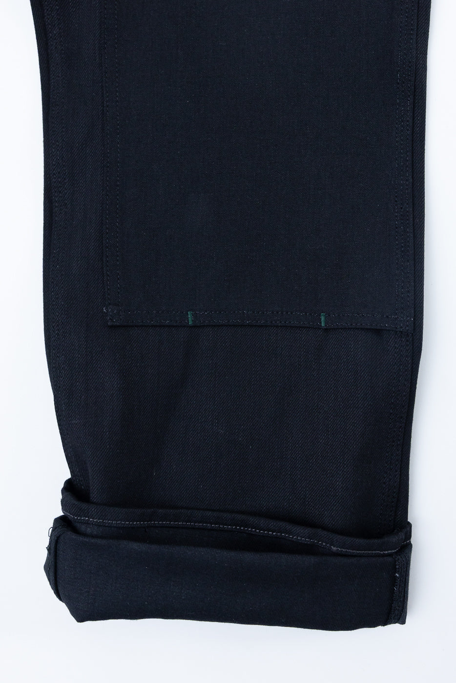 Close-up front view of dark selvedge denim pants with rolled hem and green stitching detail