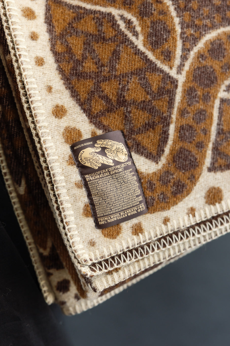 folded brown and beige blanket on a white background with custom artwork tag in lower left corner, very close up of tag with hands sigil 