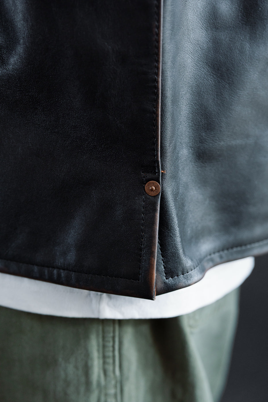 Close-up of the side hem and button detail on a black leather jacket worn by a male model.